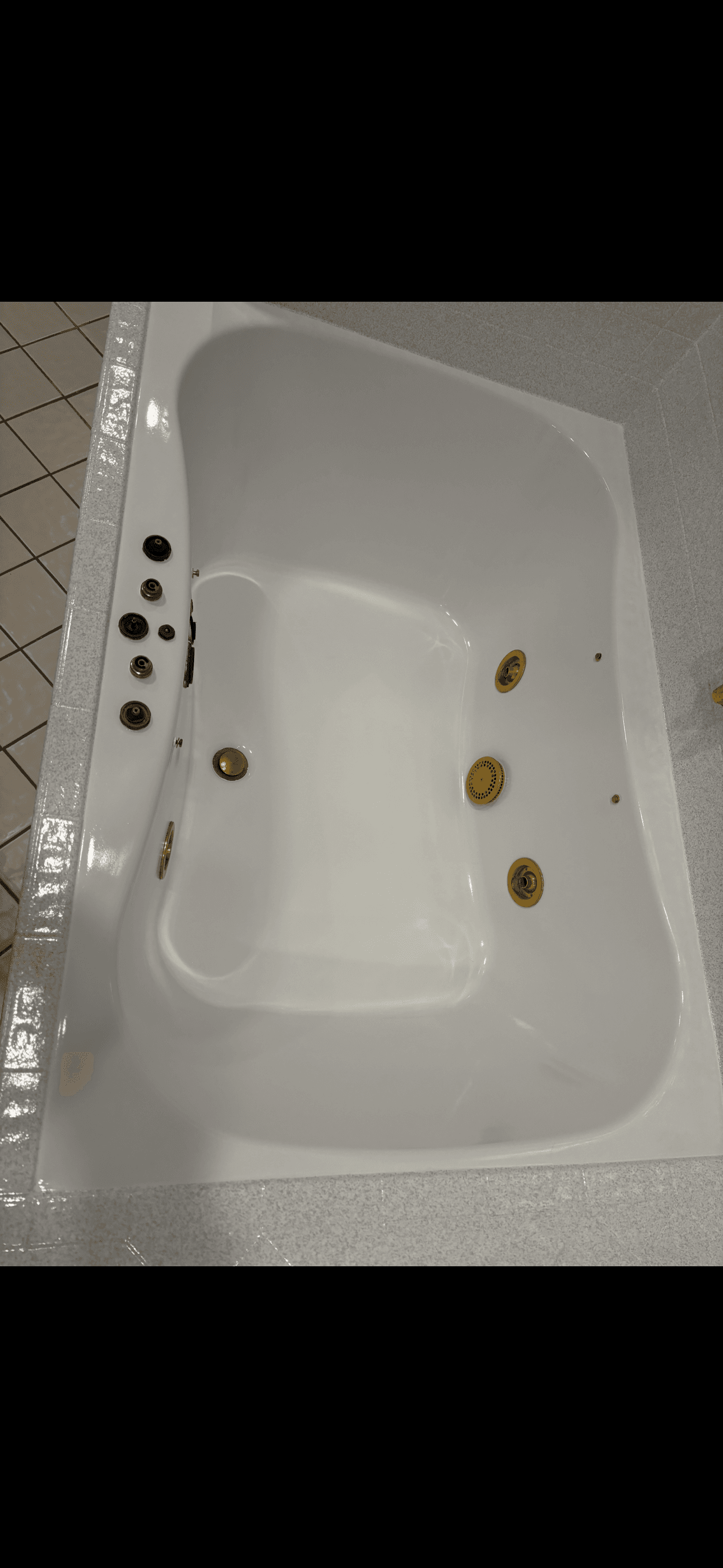 Empty white whirlpool bathtub featuring gold-colored jets and black controls on a tiled surround.
