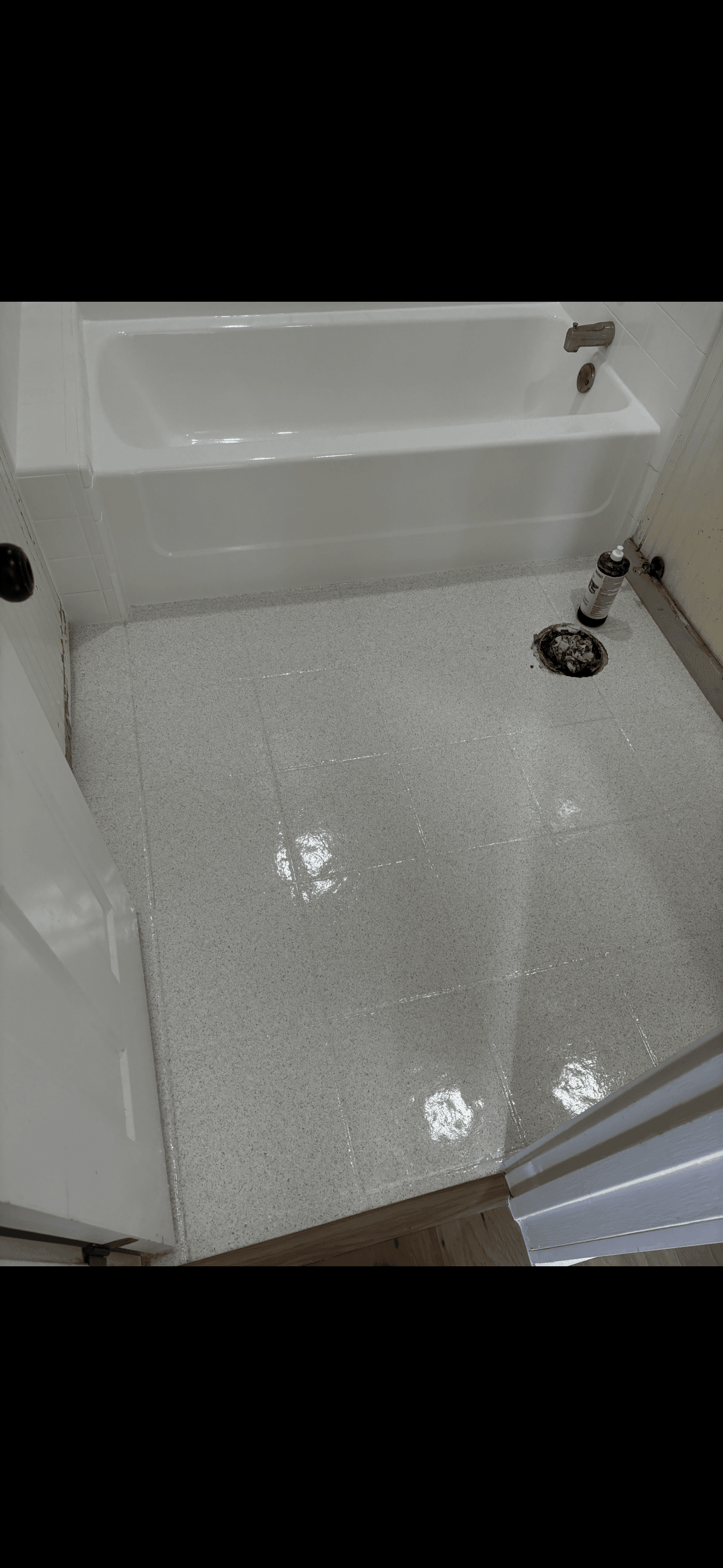 Glossy light gray speckled bathroom floor tiles next to a white bathtub and toilet flange.