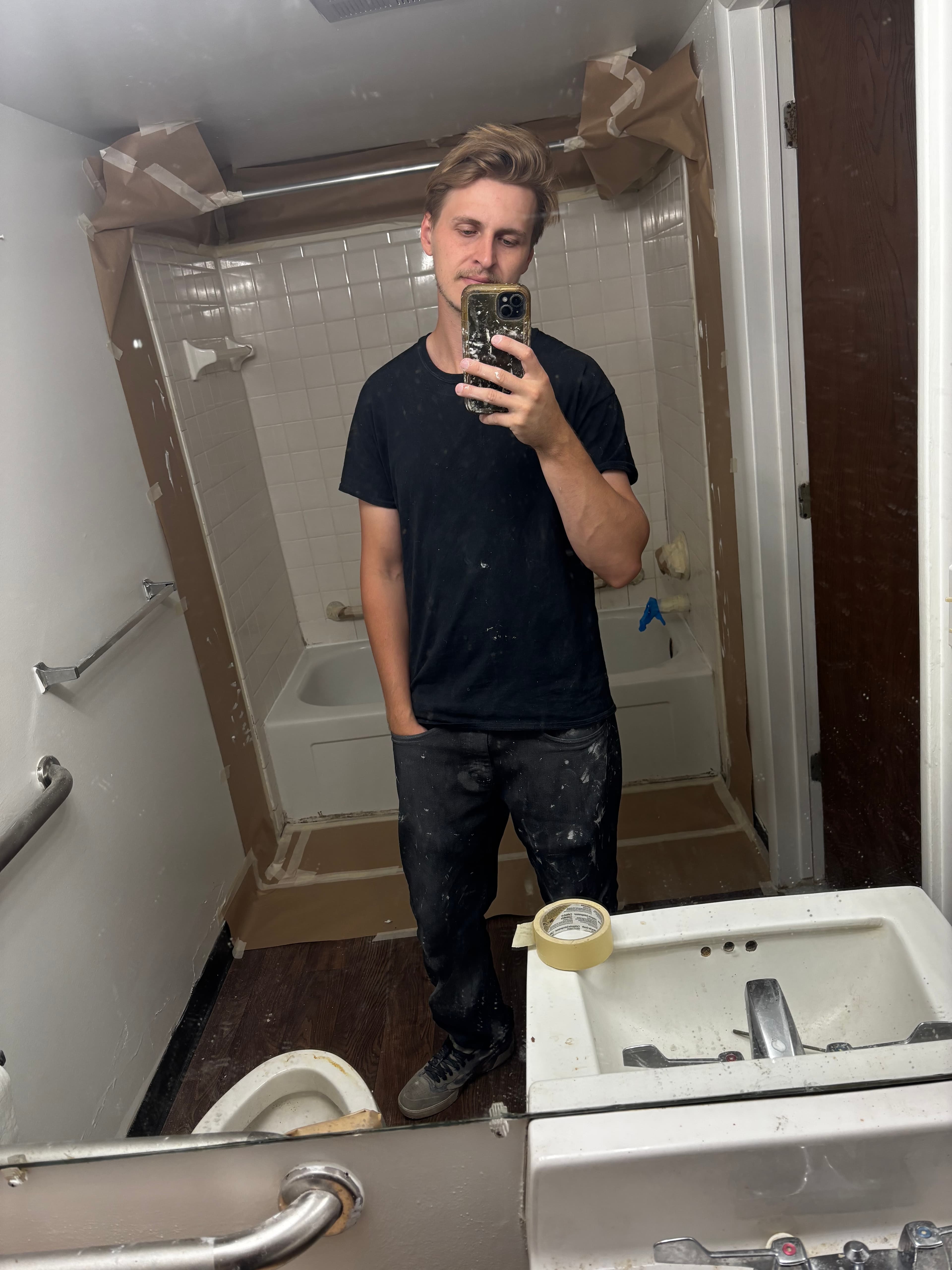 Man in paint-splattered black clothing takes a mirror selfie in a bathroom under renovation.