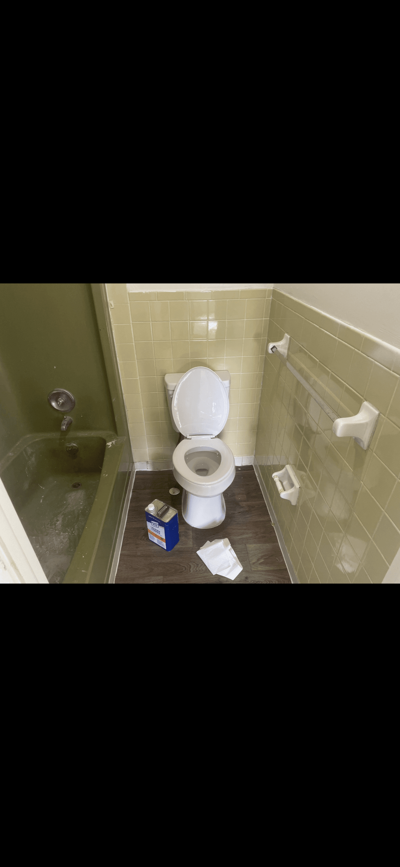 White toilet in a green tiled bathroom with a blue chemical can and crumpled paper.