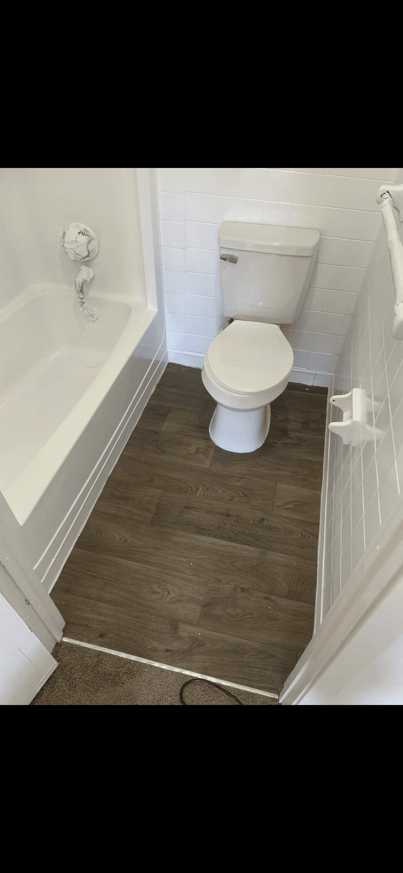 Small bathroom with white bathtub, toilet, tiled walls, and dark wood-look laminate flooring.